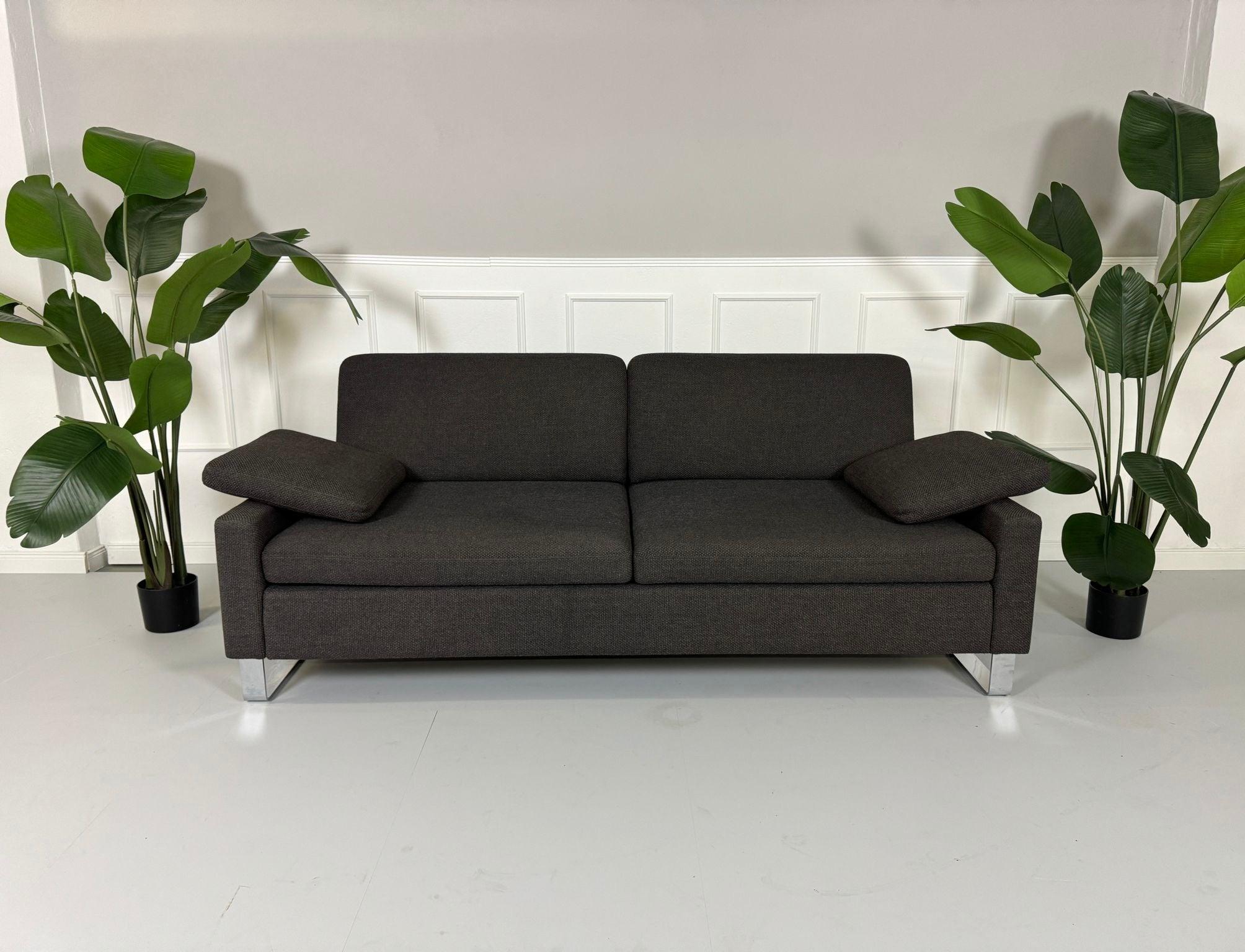 Gebrauchtes grau braunes Brühl Alba Stoff Sofa vor einer Wand, stilvoll und modern eingerichtet.