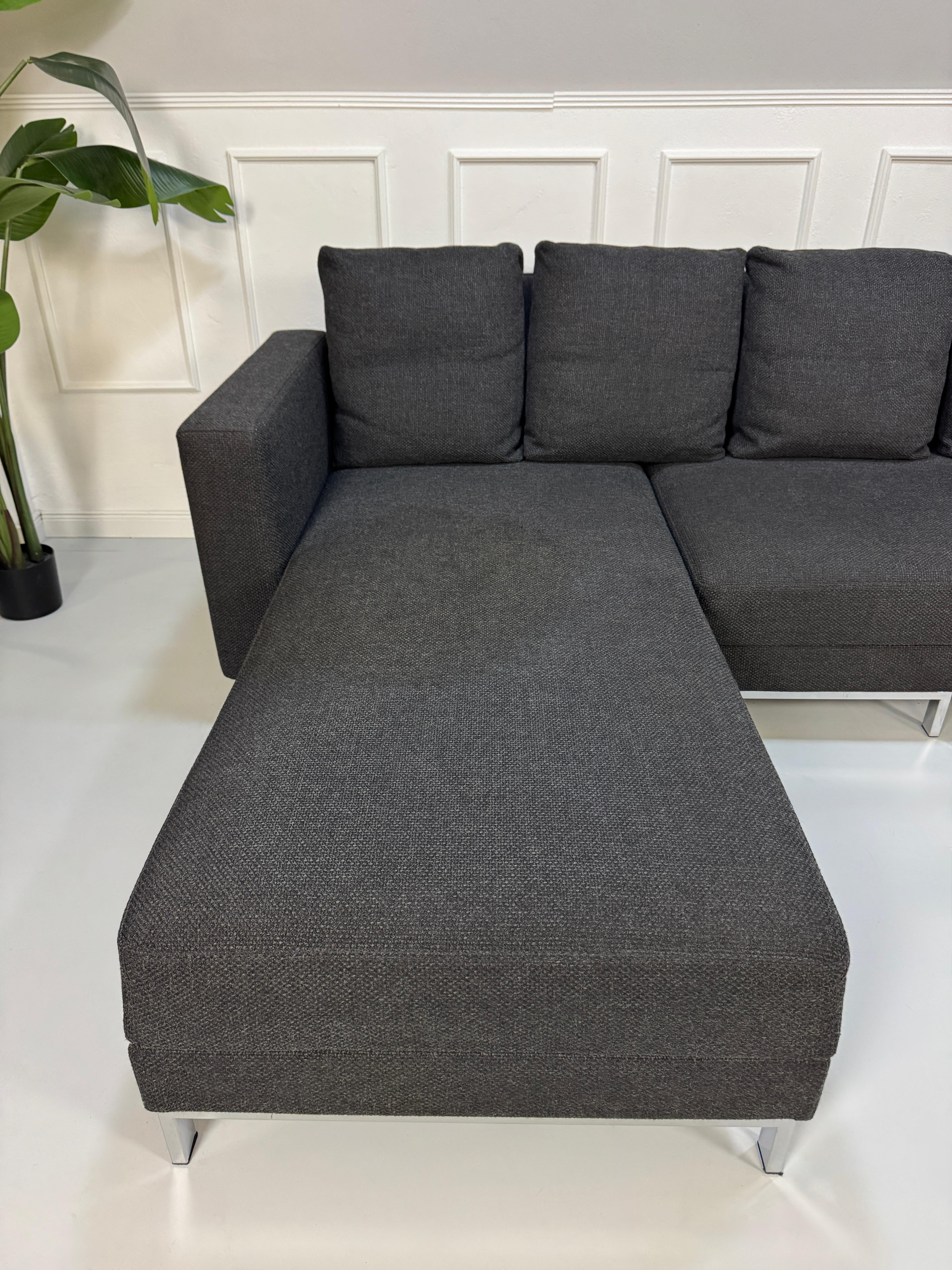 Gebrauchtes graues Brühl Fourtwo Designer Stoff Sofa vor einer Wand, stilvoll und modern eingerichtet.
