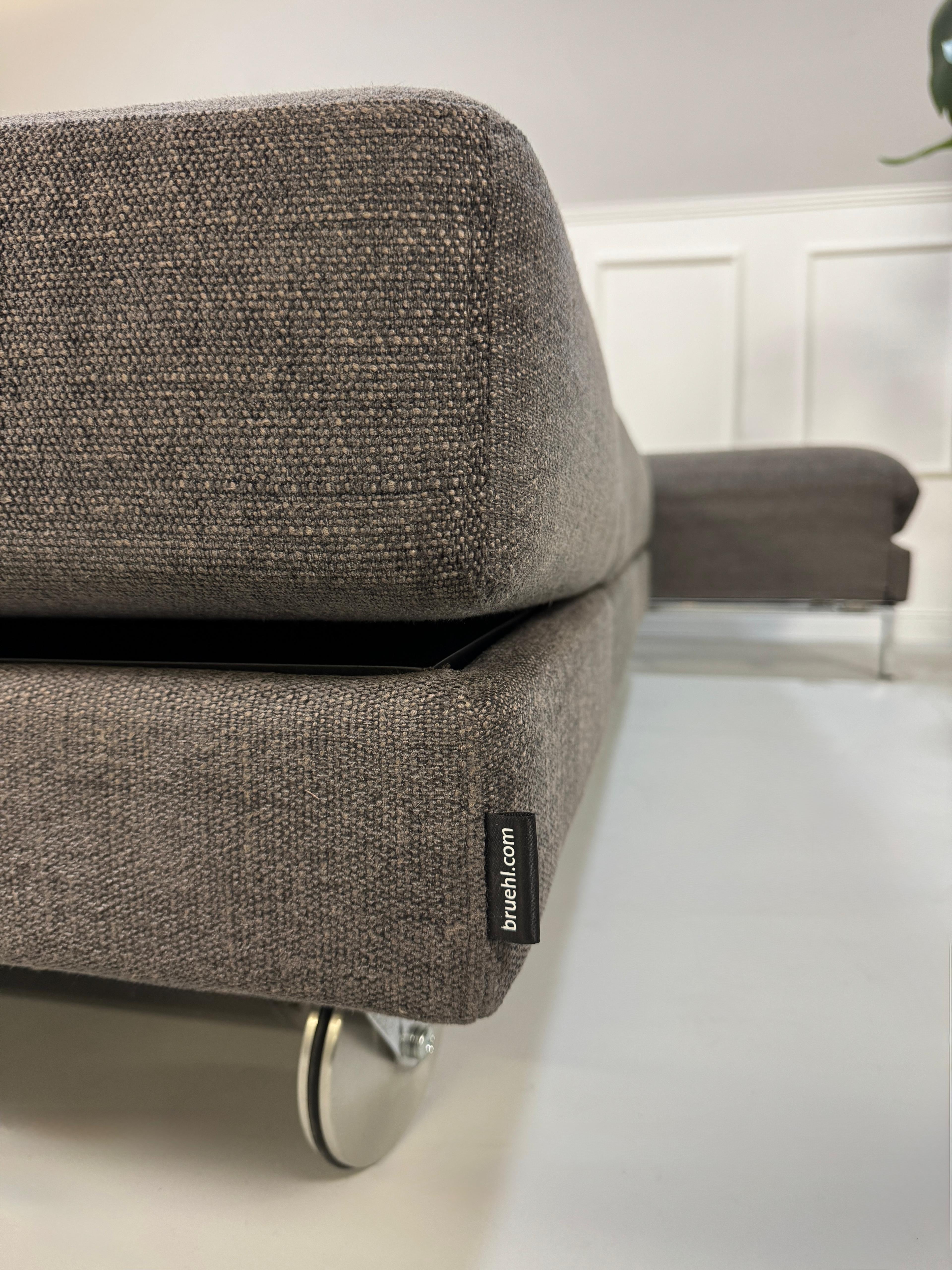 Gebrauchtes grau braunes Brühl Fourtwo Designer Stoff Sofa vor einer Wand, stilvoll und modern eingerichtet.