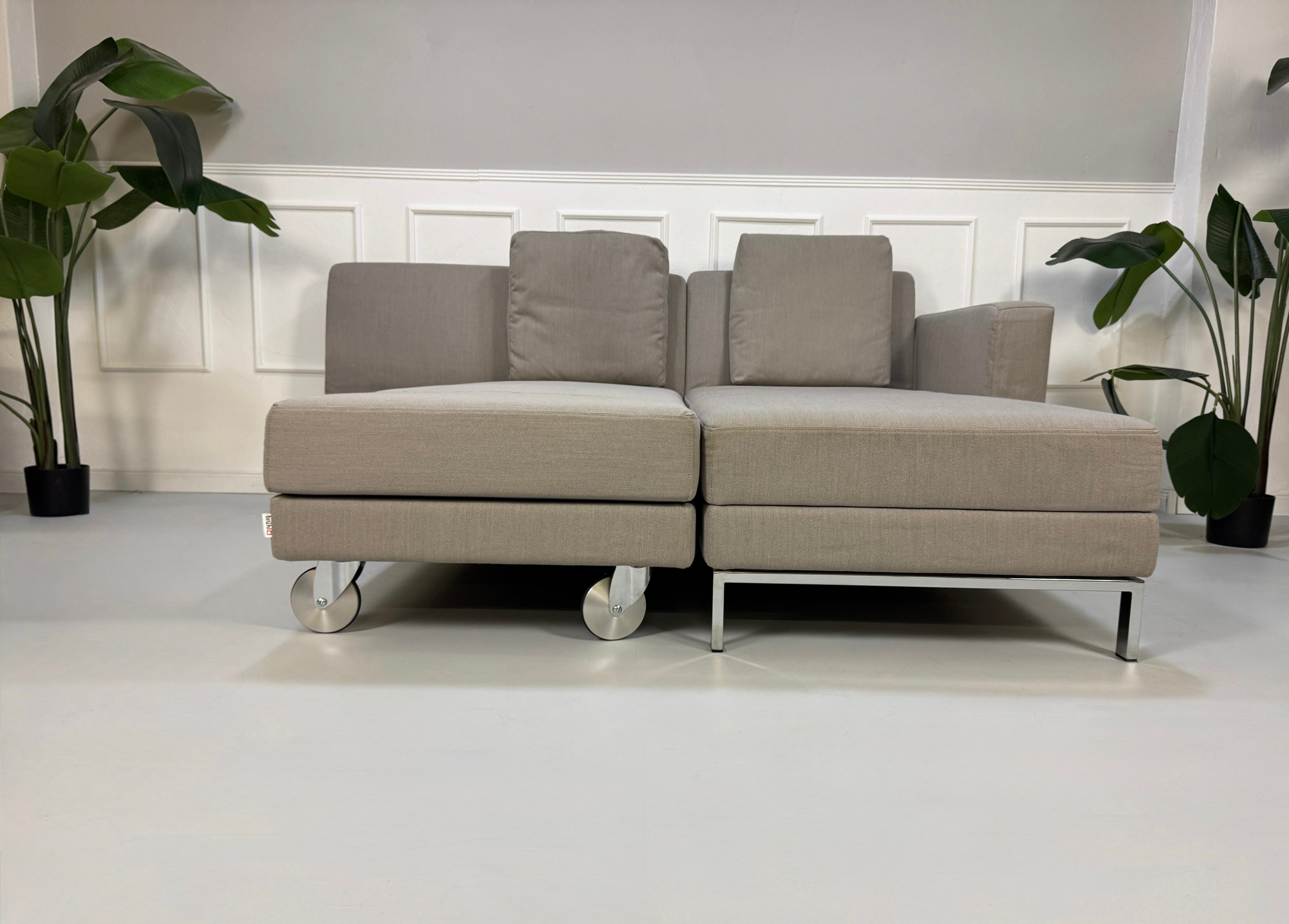 Gebrauchtes graues Brühl Fourtwo Designer Stoff Sofa vor einer Wand, stilvoll und modern eingerichtet.