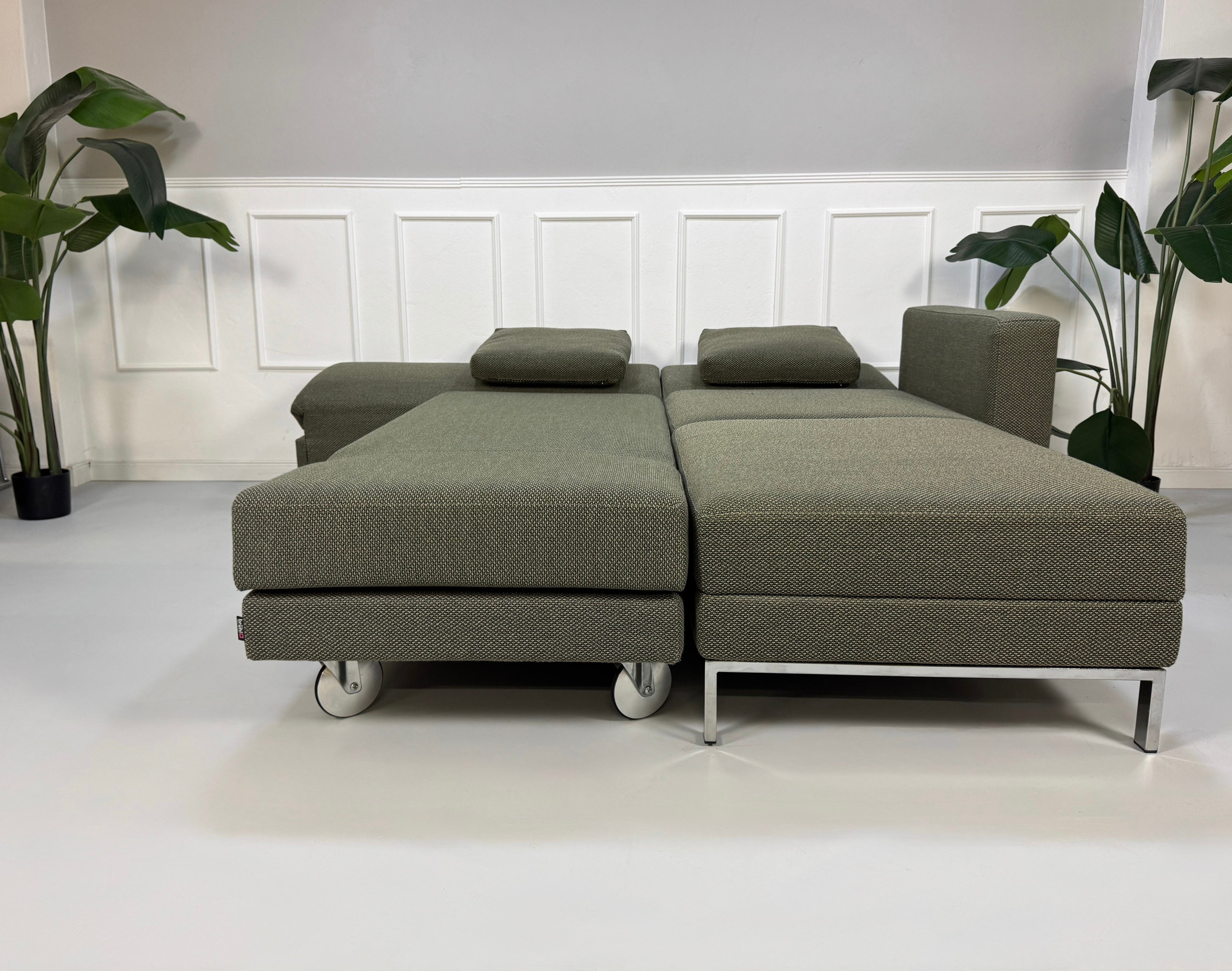 Grau Grünes Brühl Fourtwo Designer Stoff Sofa vor einer Wand, stilvoll und modern eingerichtet.