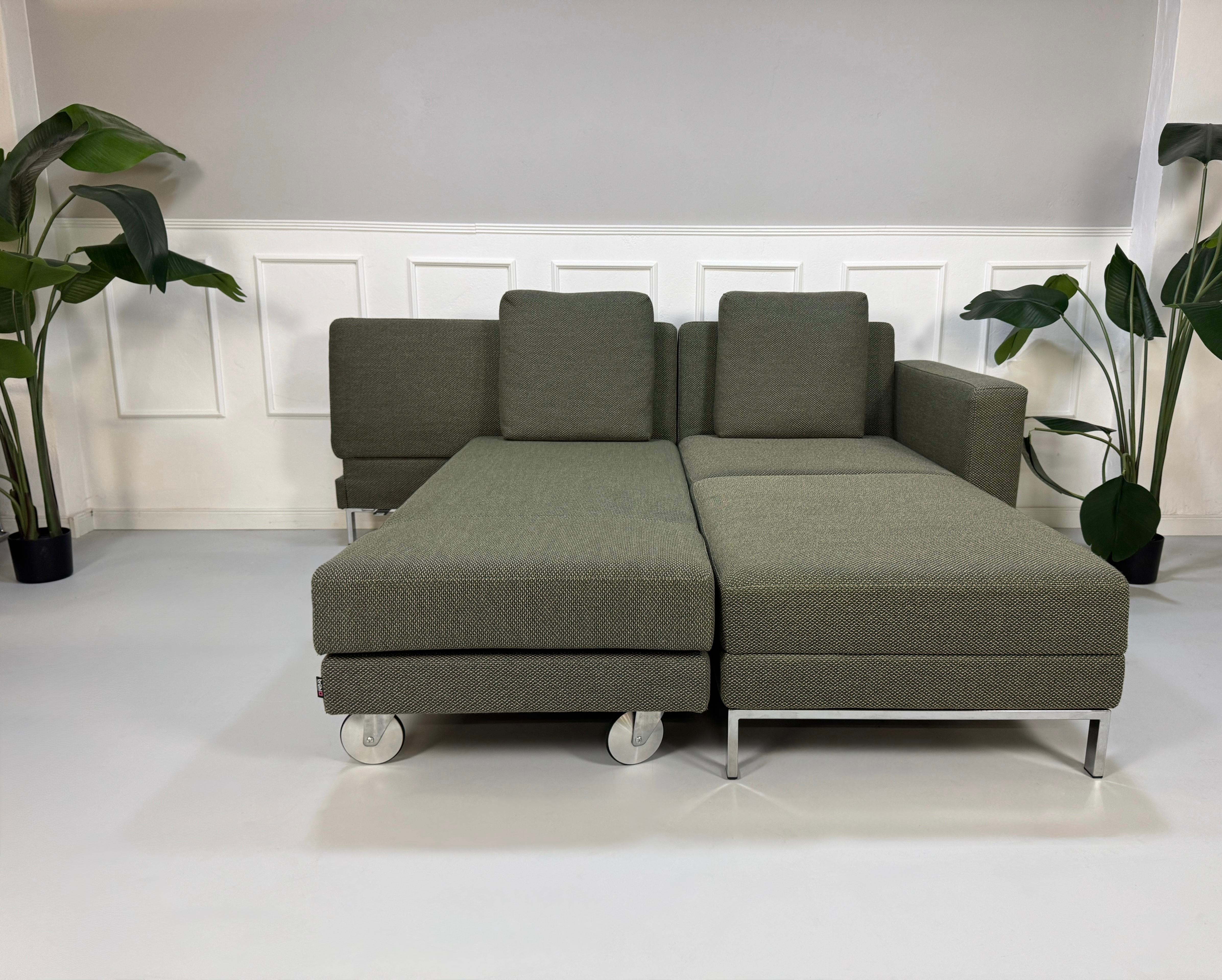 Grau Grünes Brühl Fourtwo Designer Stoff Sofa vor einer Wand, stilvoll und modern eingerichtet.