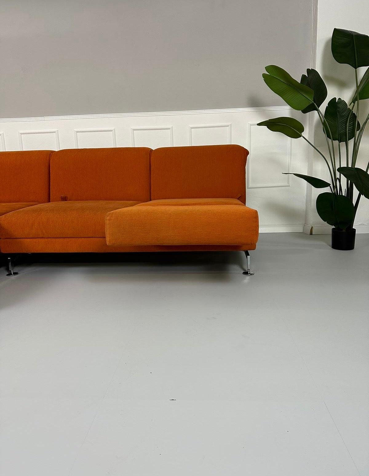 Gebrauchtes Brühl Moule Ecksofa Stoff in der Farbe Orange vor einer Wand, stilvoll und modern eingerichtet.