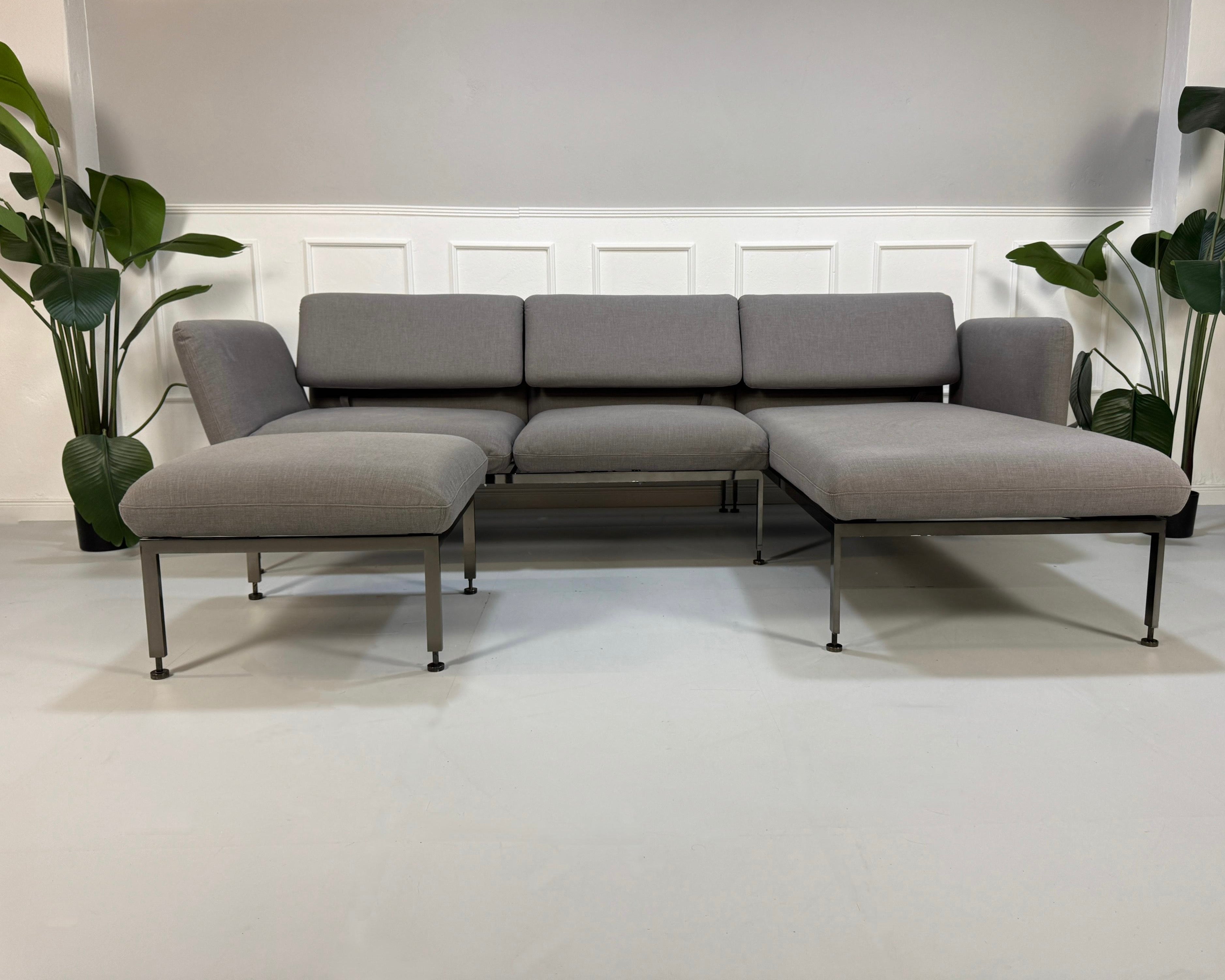 Gebrauchtes graues Brühl Roro Designer Stoff Sofa mit Hocker vor einer Wand, stilvoll und modern eingerichtet.
