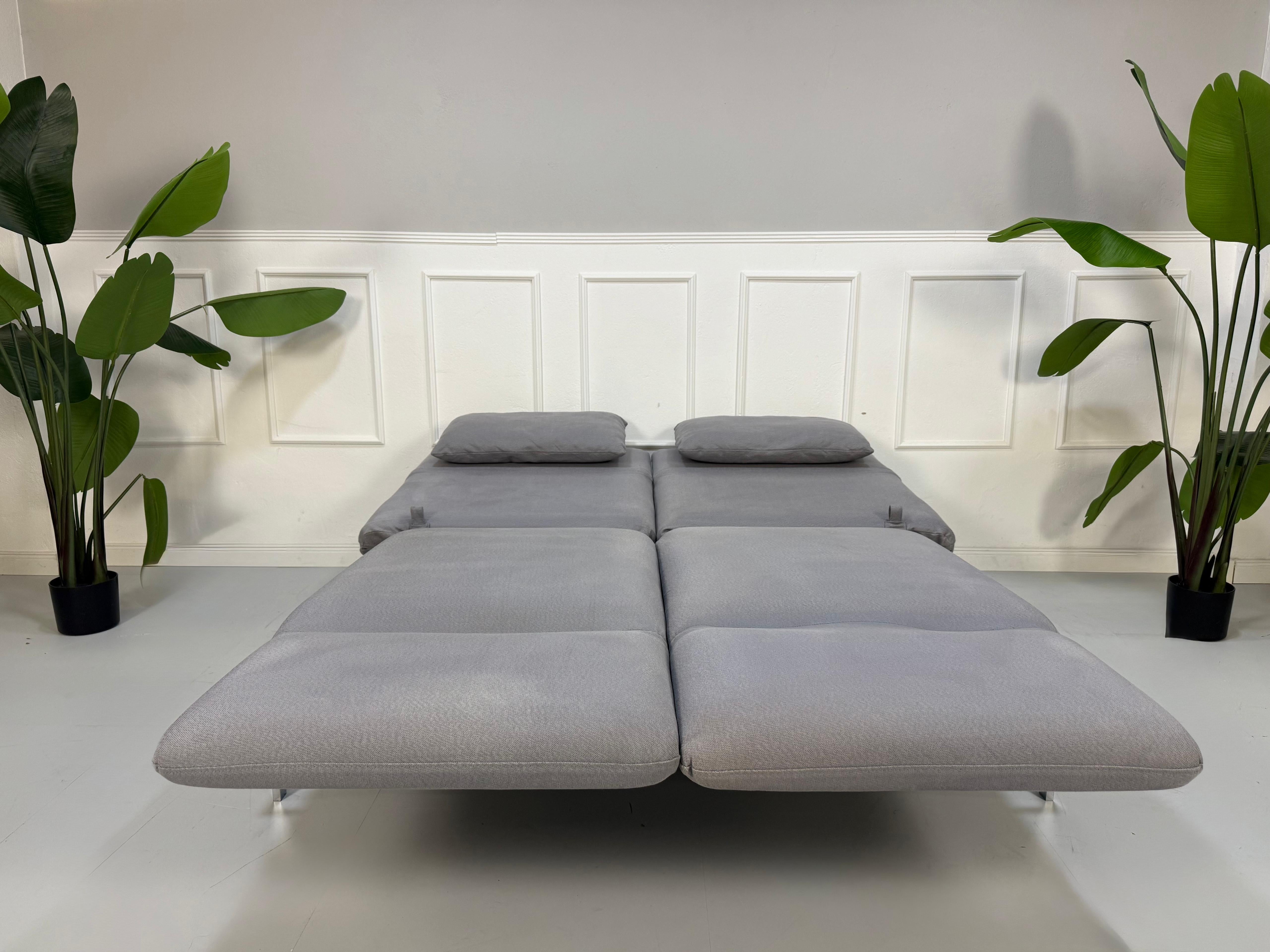 Erneuertes Brühl Roro Designer Stoff Sofa in der Farbe Grau vor einer Wand, stilvoll und modern eingerichtet.