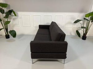 Gebrauchtes braun graues Brühl Tomo Designer Stoff Sofa mit Hocker vor einer Wand, stilvoll und modern eingerichtet.