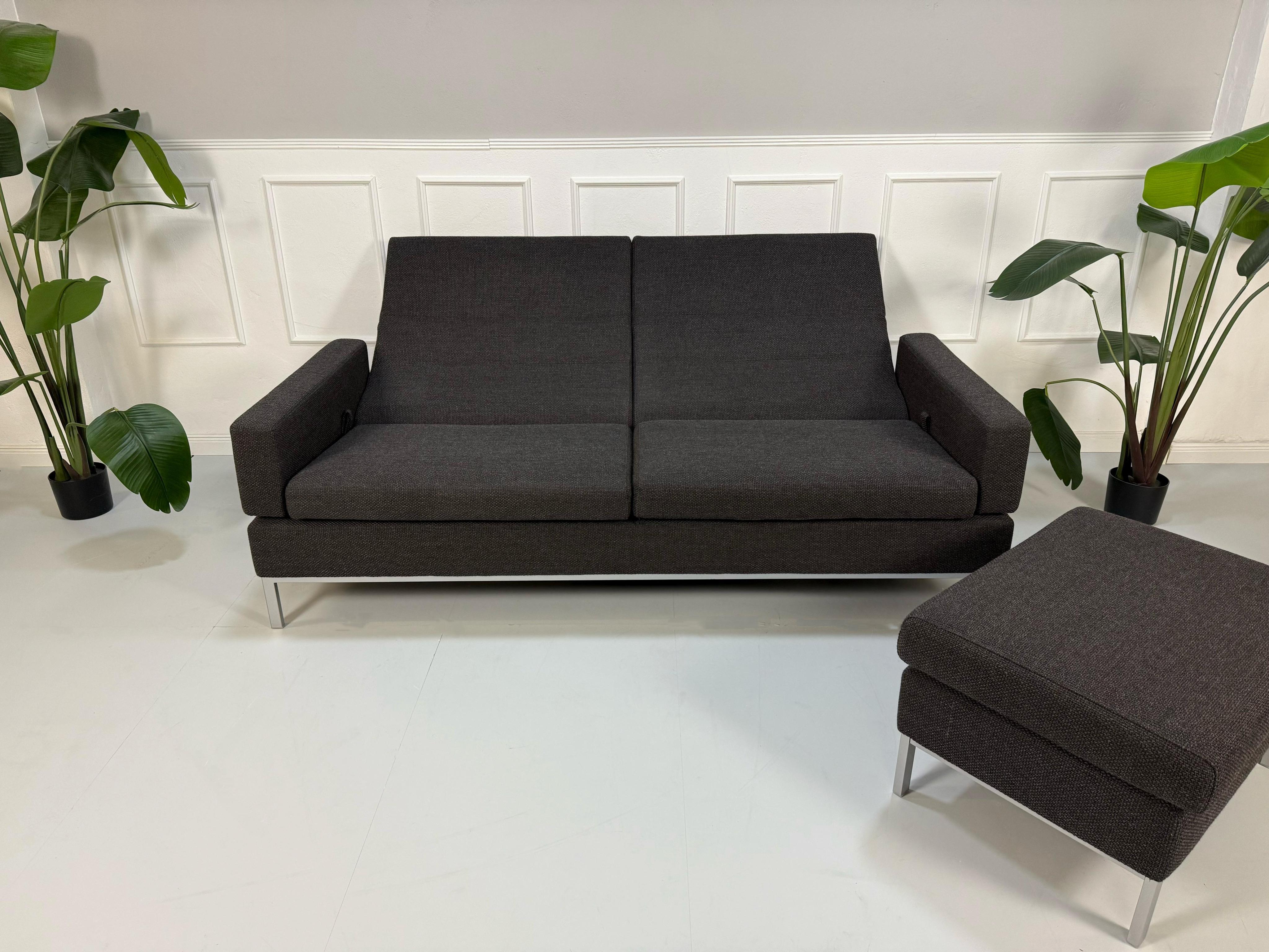 Gebrauchtes braun graues Brühl Tomo Designer Stoff Sofa mit Hocker vor einer Wand, stilvoll und modern eingerichtet.