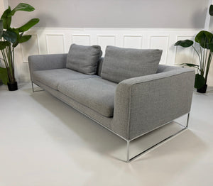 Erneuertes graues COR Mell Designer Stoff Sofa vor einer Wand, stilvoll und modern eingerichtet.