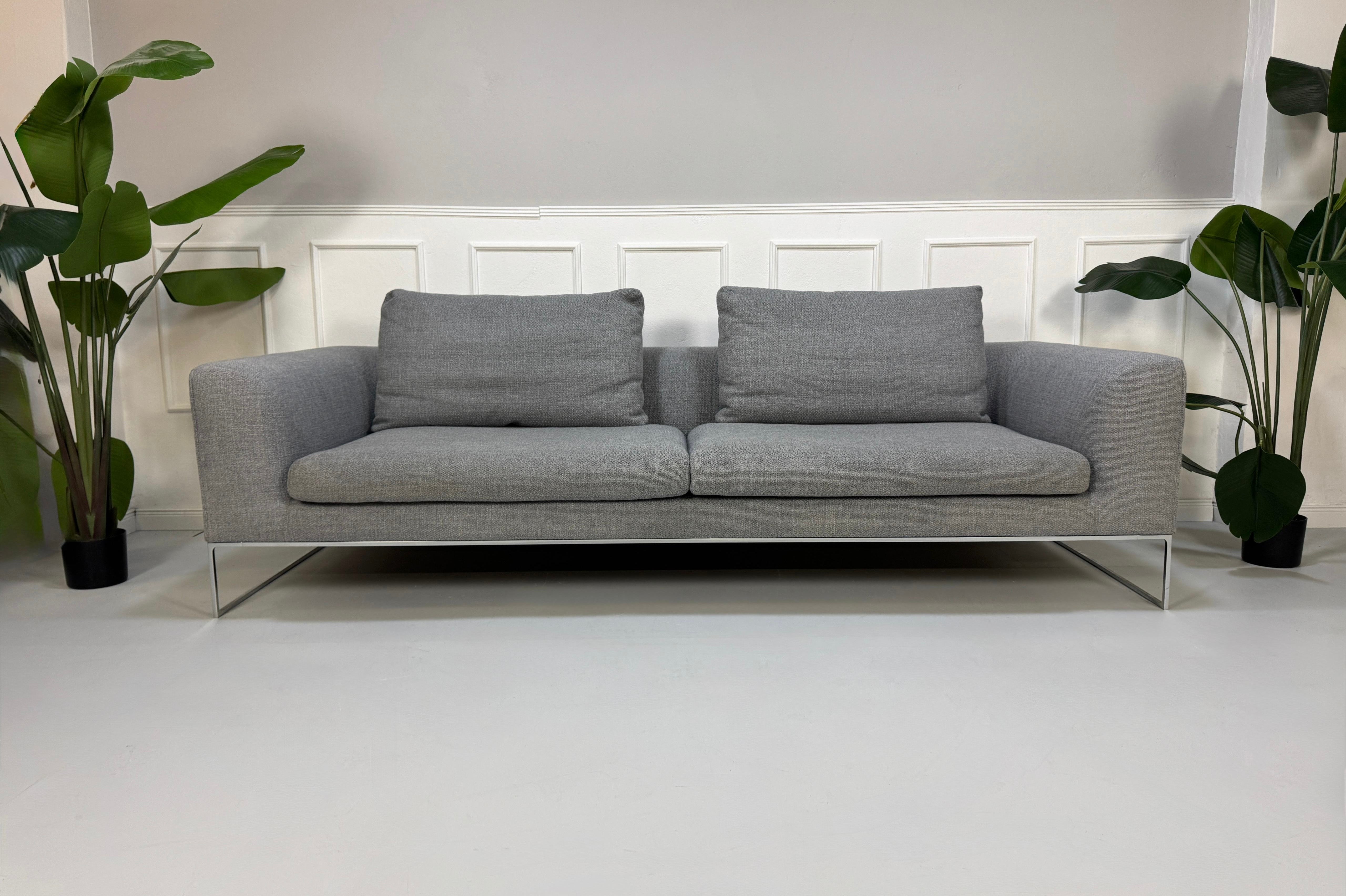 Erneuertes graues COR Mell Designer Stoff Sofa vor einer Wand, stilvoll und modern eingerichtet.