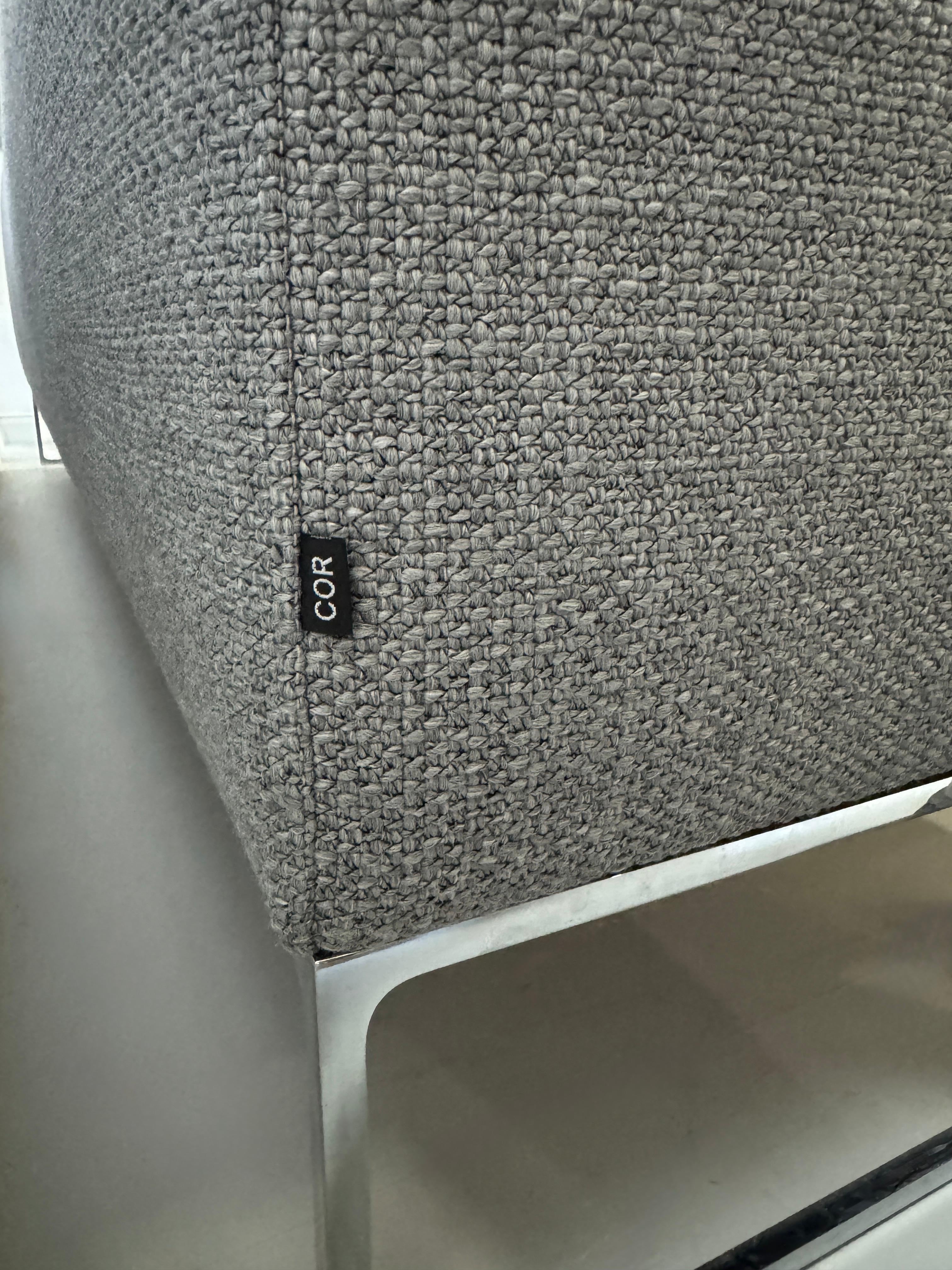 Erneuertes graues COR Mell Designer Stoff Sofa vor einer Wand, stilvoll und modern eingerichtet.