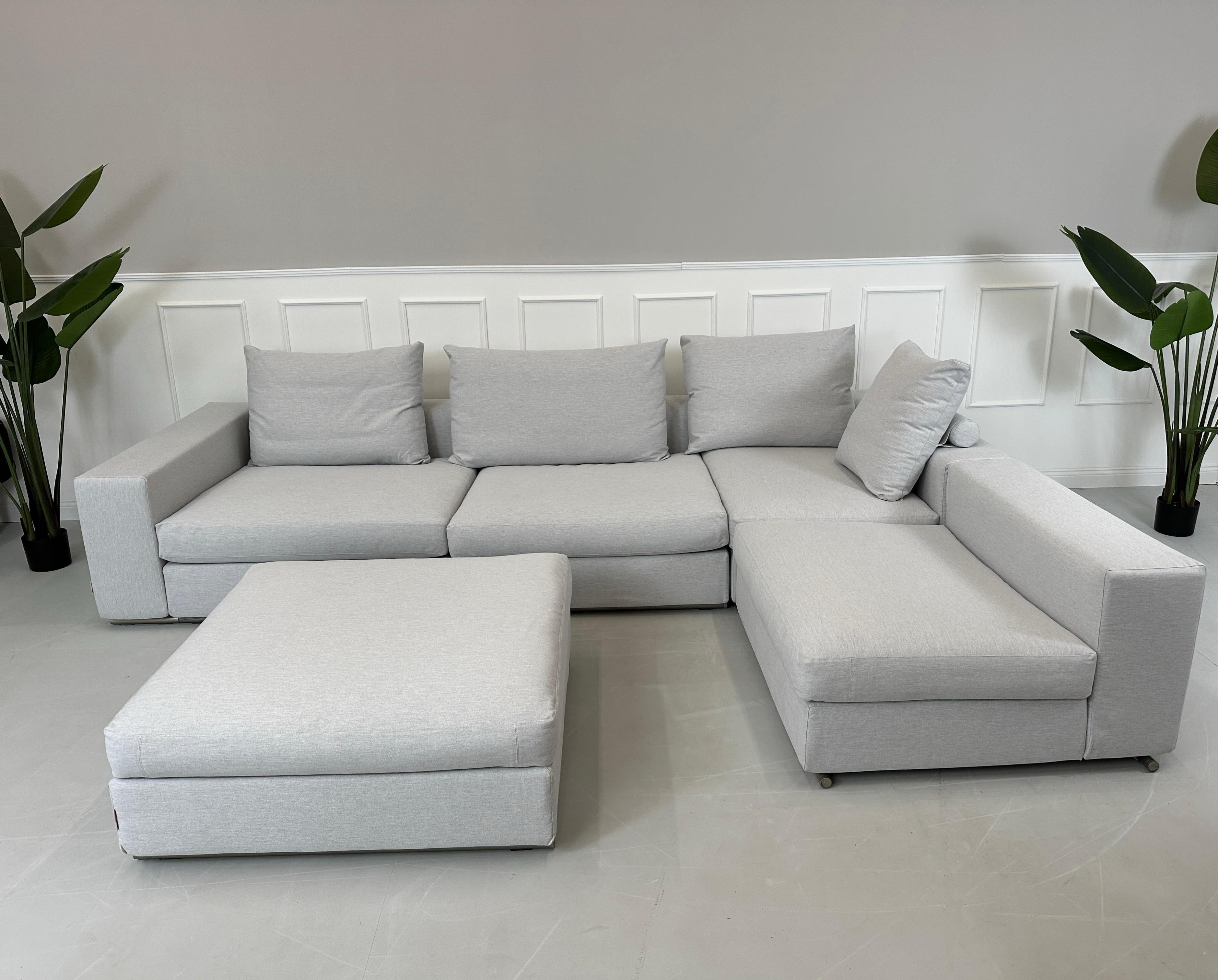 Gebrauchtes graues Flexform Groundpiece Designer Stoff Sofa mit Hocker vor einer Wand, stilvoll und modern eingerichtet.