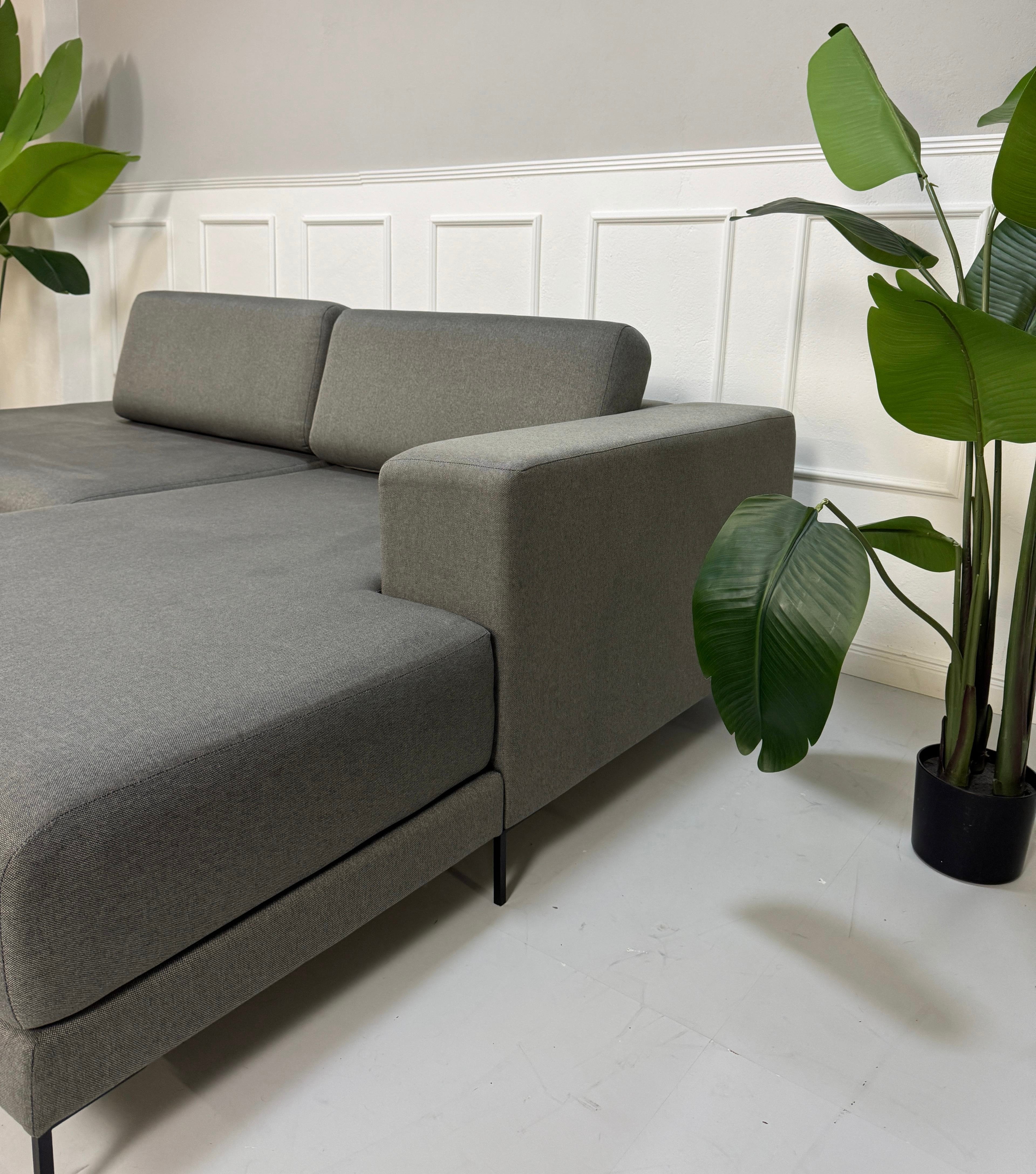 Gebrauchtes graues MYCS Tyme Designer Stoff Sofa vor einer Wand, stilvoll und modern eingerichtet.