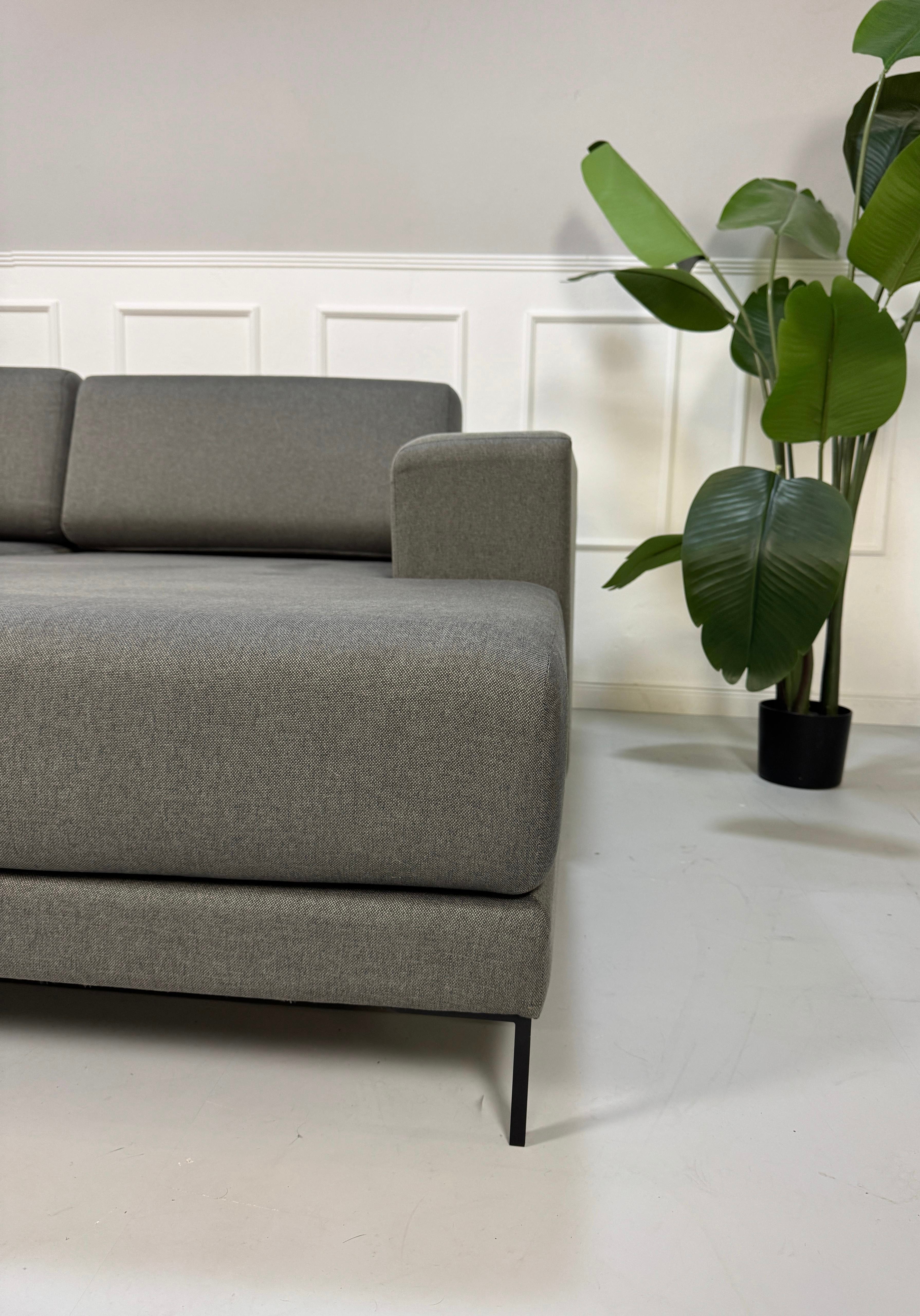 Gebrauchtes graues MYCS Tyme Designer Stoff Sofa vor einer Wand, stilvoll und modern eingerichtet.