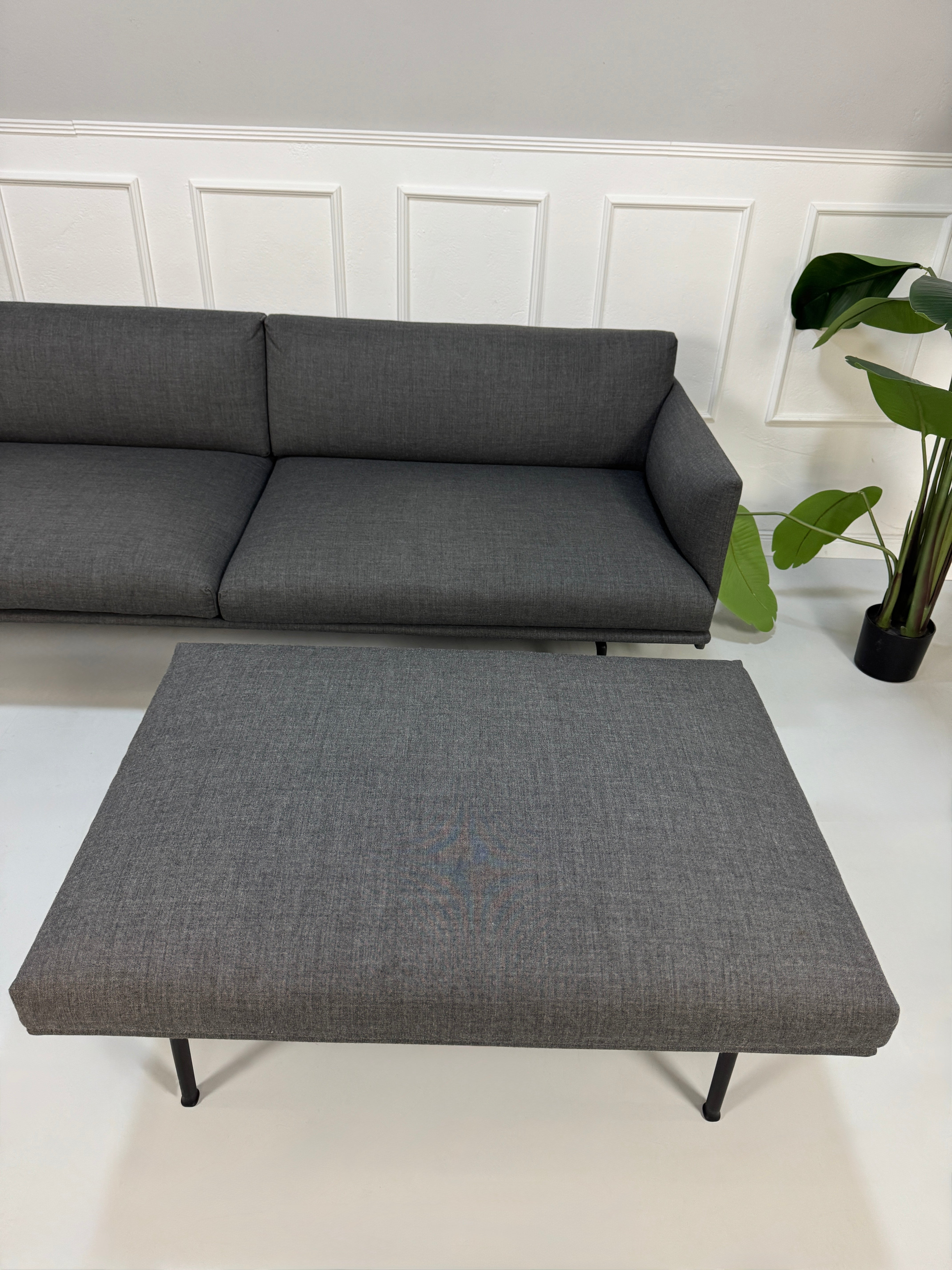 Gebrauchtes graues Muuto Outline Designer Stoff Sofa mit einem Hocker vor einer Wand, stilvoll und modern eingerichtet.