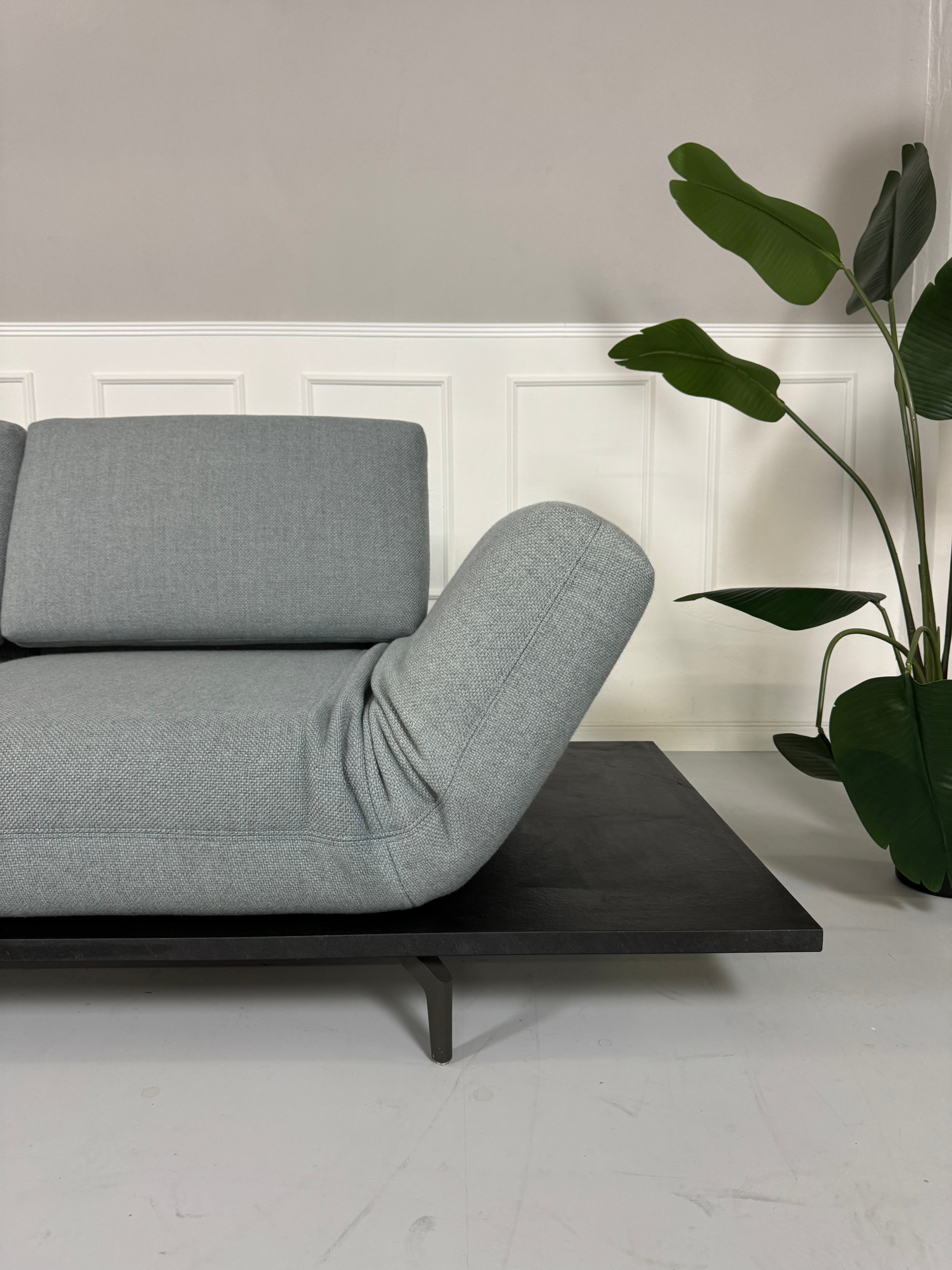 Gebrauchtes grau blaues Rolf Benz Aura Designer Stoff Sofa vor einer Wand, stilvoll und modern eingerichtet.