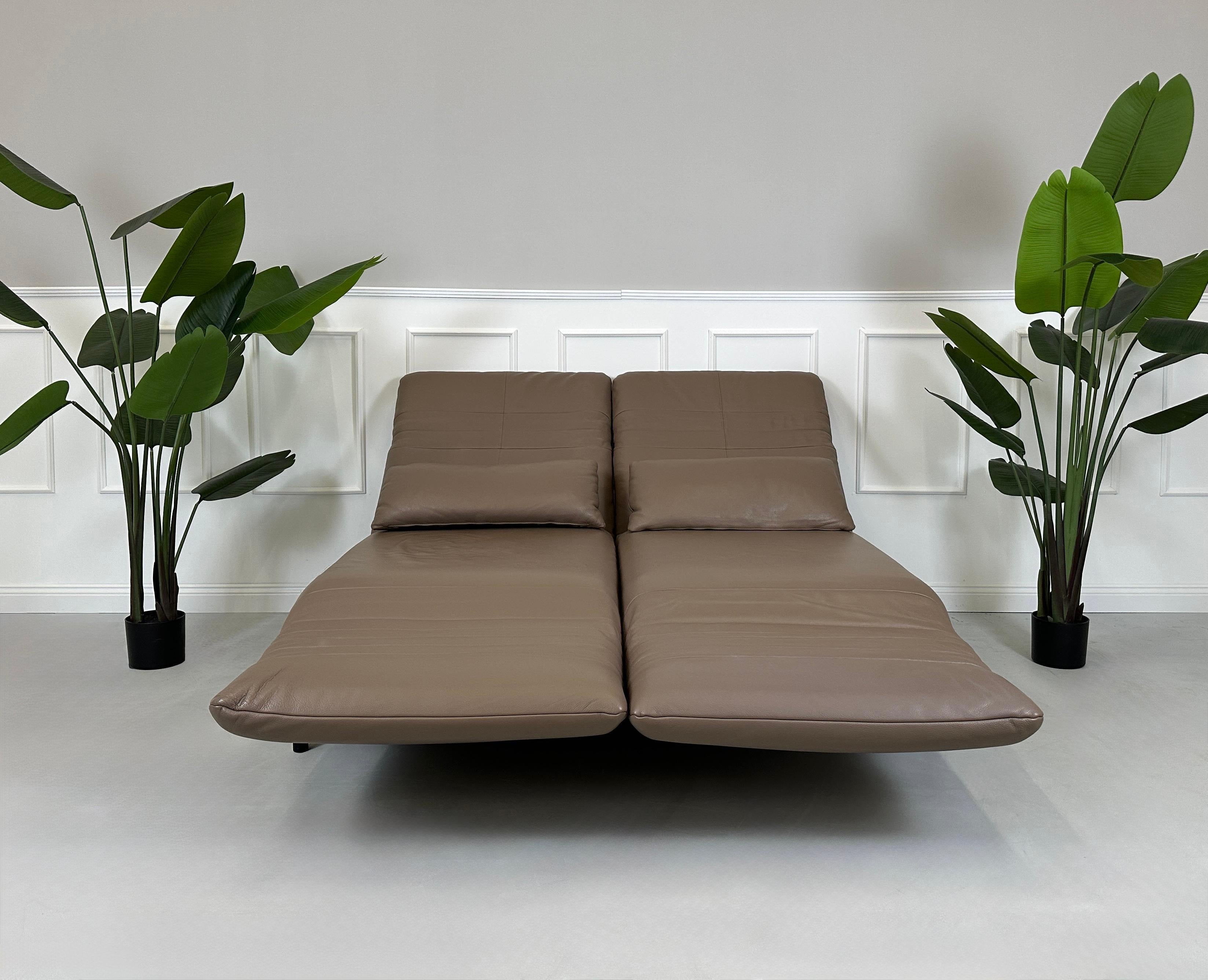 Gebrauchtes braunes Rolf Benz Plura Designer Leder Sofa mit Hocker vor einer Wand, stilvoll und modern eingerichtet.