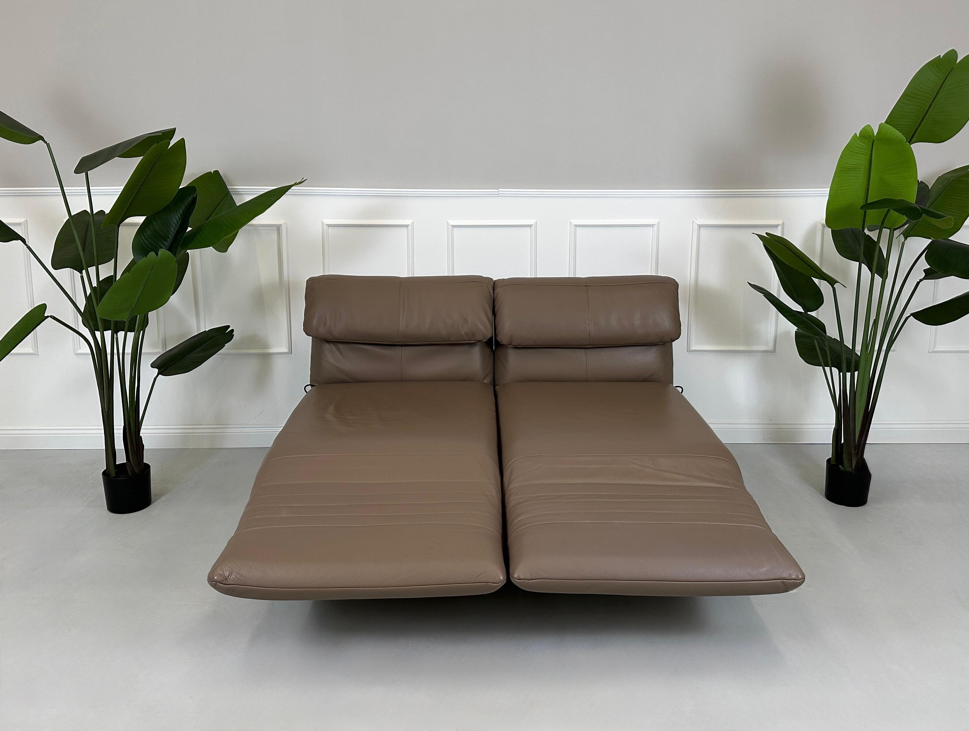 Gebrauchtes braunes Rolf Benz Plura Designer Leder Sofa mit Hocker vor einer Wand, stilvoll und modern eingerichtet.