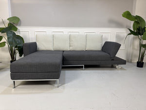 Gebrauchtes graues Brühl Fourtwo Designer Stoff Ecksofa vor einer Wand, stilvoll und modern eingerichtet.