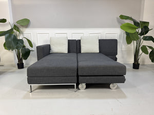 Gebrauchtes graues Brühl Fourtwo Designer Stoff Ecksofa vor einer Wand, stilvoll und modern eingerichtet.
