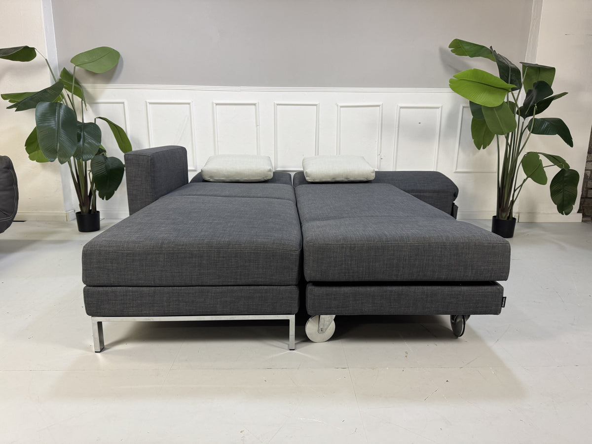 Gebrauchtes graues Brühl Fourtwo Designer Stoff Ecksofa vor einer Wand, stilvoll und modern eingerichtet.
